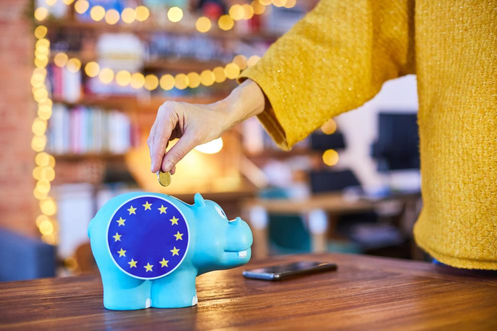 Krepitev dolgoročnega proračuna EU (Foto: oseba v rumeni majici daje kovanec v hranilnik v obliki modrega prašička z nalepko zastave EU)
