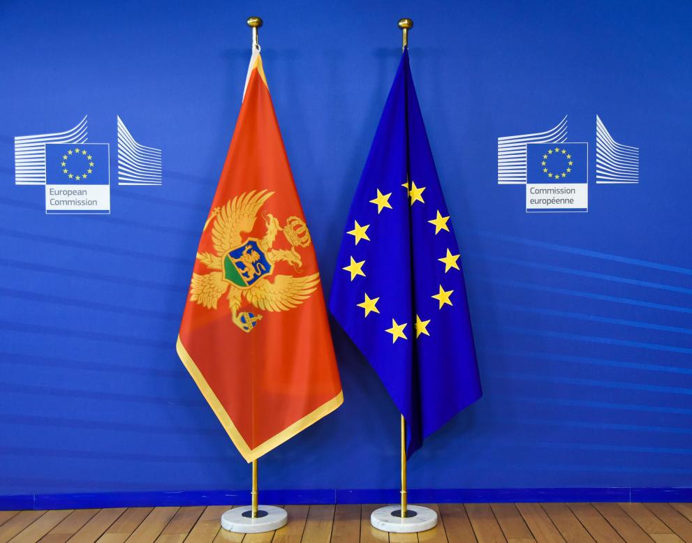 Črna gora vključena v program EU4Health (zastavi Črne gore in EU visita na dveh ločenih stojalih pred modro steno z logotipi Evropske komisije