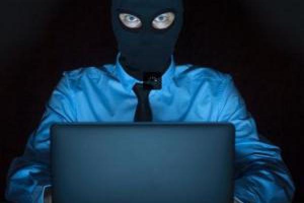 Kibernetska varnost (Foto: moški z vlomilsko masko, črno masko z izrezanimi očmi, sedi pred odprtim notesnikom in grozeče gleda v svet)